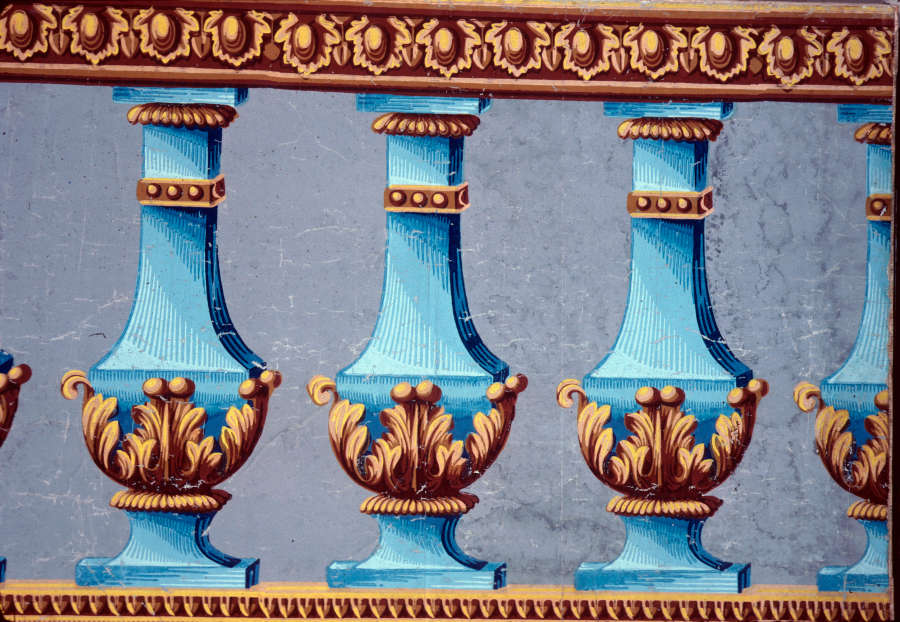 Print of repeating blue vases with a floral, geometric border above and thin lined border below. Yellow dotted embellishments are along its neck and raised leaf motifs around its base.