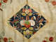 A close up of a diamond-shaped embroidered panel with intricate red, green, and beige designs of curling vines and delicate blossoms against a deep blue background on cream-colored fabric.