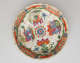 A white saucer with green, red, blue, pink, yellow, and gilded decorations; separated into four main vignites, two animals and two tables and vessels containing floral elements.
