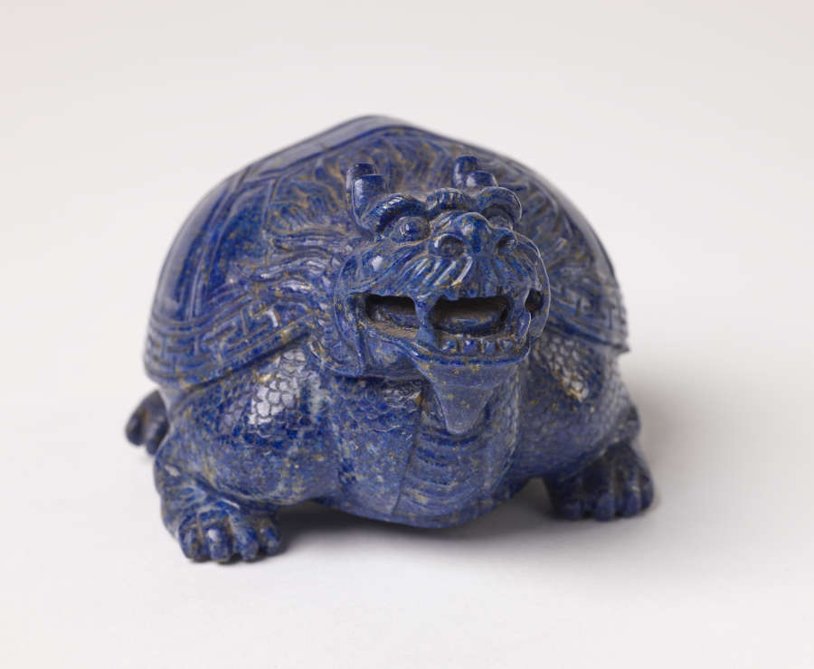 A head-on view of a blue sculpture of a smiling horned tortoise, whose surface is covered with carved geometric patterns.
