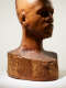 Dramatically lit, three quarter view of a wooden-carved bust depicting a figure with afrocentric facial features with the top of the head out of frame. “Elizabeth Prophet” is carved into the side.