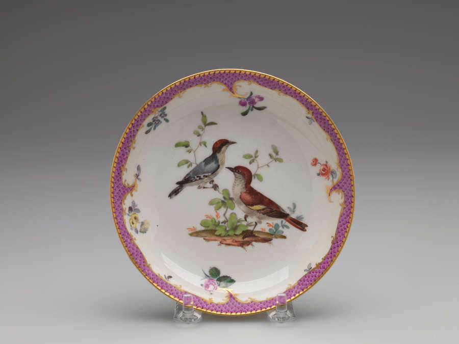 A white plate with a gilded and pink edge and floral patterns. Center design features two birds perched on leafy branches.