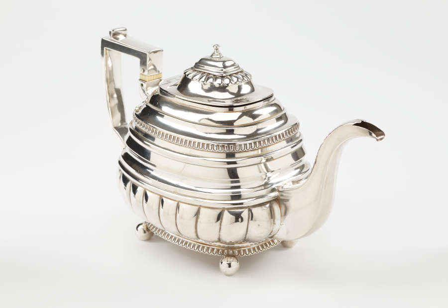 A silver teapot with a decorative angular handle, a rounded square body and spout with sculptural decorations.