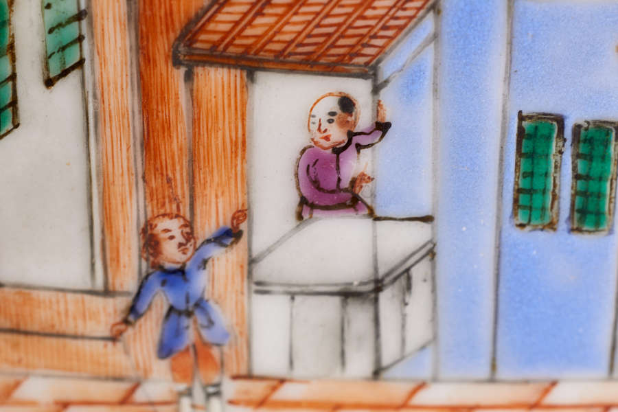 A close up of a painted ceramic depicting a figure in blue standing on an orange brick street near a doorway and a figure dressed in purple at a counter.