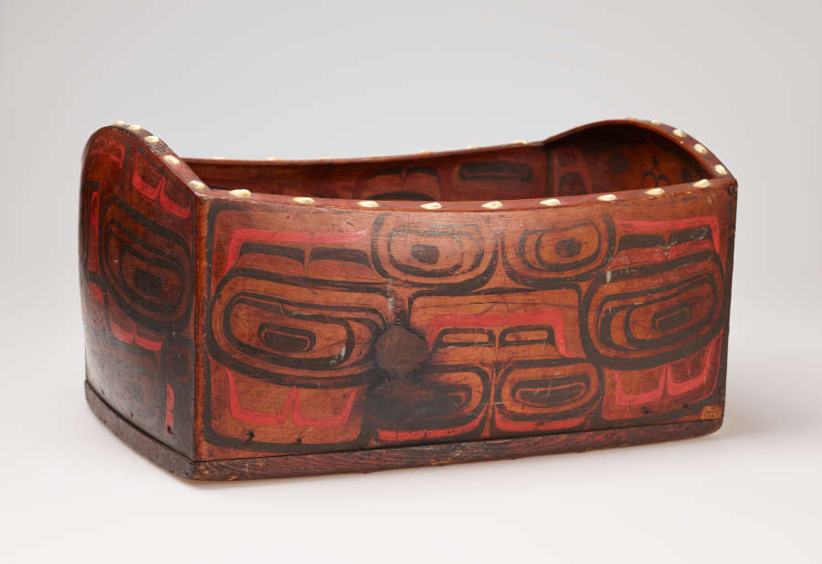 Three-quarter view of a wooden box with red and black graphic motifs resembling creatures and faces. The box’s rim undulates and has a line of large white dots decorating it.