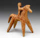 Terracotta figurine of the simplified form of a horse with rider, decorated with red painted patterns. The patterns emphasize the horse’s elongated form and the rider’s posture, gripping the horse.
