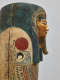 Angled view of a coffin with a gilded face, blue-black headdress and beard. A red sun above a falcon is painted on the shoulder and intricate patterns decorate the chest.