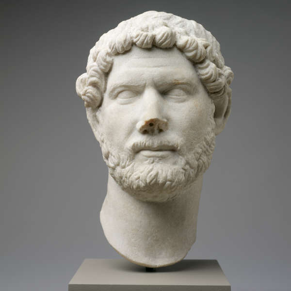 Marble bust from the neck up of a realistically carved bearded man with loose curly hair and furrowed brows resting on a pedestal against a gray backdrop.