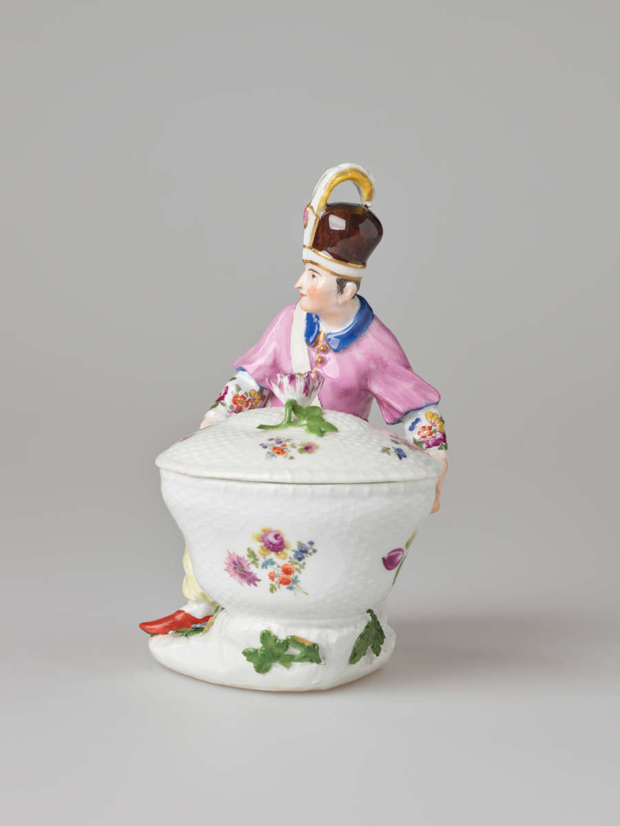 A sculptural figure kneeling next to a basket. Figure wears bright, elaborate clothing with a hat with a feather. There are floral decorations on the figure’s clothing and basket.
