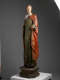 A tall, weathered wooden statue of a robed figure with a serene expression, draped in green and red garments, standing upon an octagonal base.