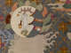 Intricate embroidery depicting a scene of a white, long eared creature holding a bucket beside a pagoda, contained within a circle surrounded by swirling clouds and flowers in a grid.