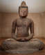 An aged weathered wooden Buddha statue sat cross-legged in meditation with a serene expression in a warmly lit room.