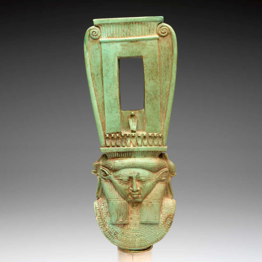 Front view of a long jade-green sculpture of a collared figure wearing a tall headdress, which has a rectangular opening and swirls on the outer edges forming a curved top.