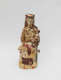 An intricately painted ceramic figure of a seated woman wearing a headdress cradling a small child in her left arm, the surface richly patterned with beige, red, creams, and browns.