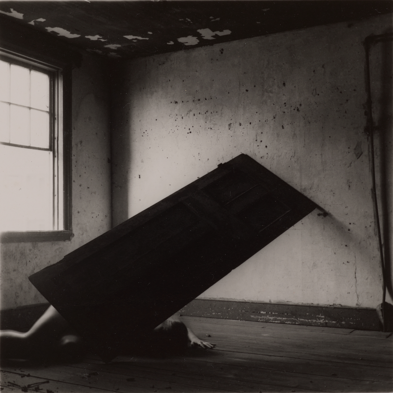 Dreamy black and white photo of a door propped precariously against a wall. A figure crouches beneath, a pale arm and leg visible. Light streams in a window at left.