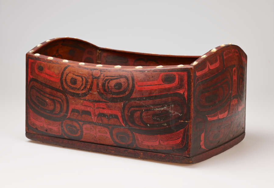 Three-quarter view of a wooden box with red and black graphic motifs resembling creatures and faces. The box’s rim undulates and has a line of large white dots decorating it.