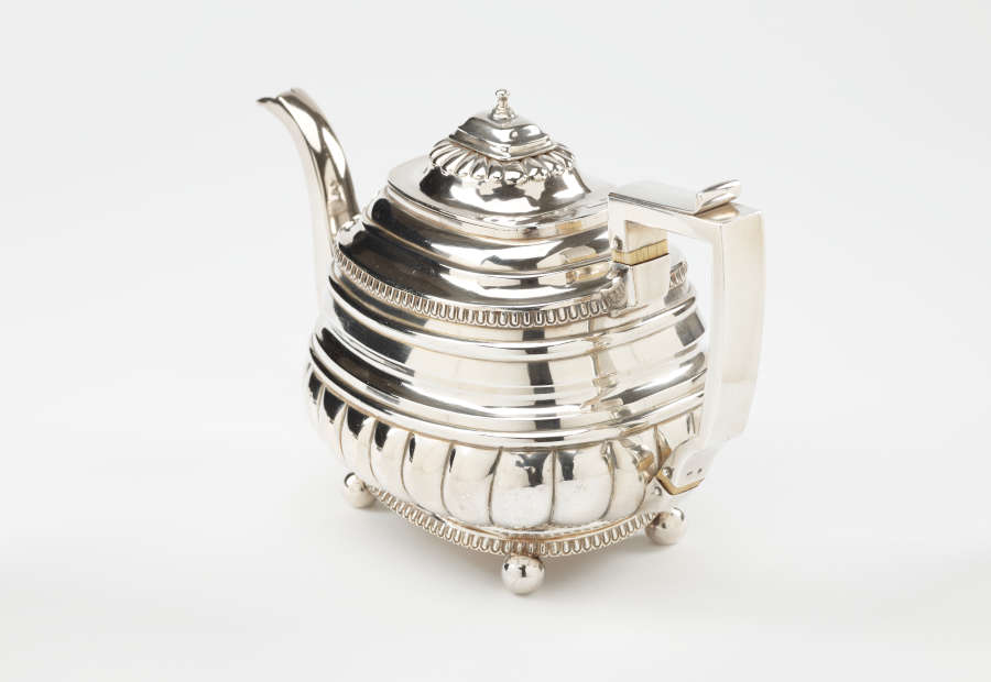 A silver teapot with a decorative angular handle, a rounded square body and spout with sculptural decorations.