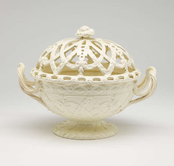 A cream-colored basket with sculptural woven handles, sculpted foot, and a lid with cutout designs.