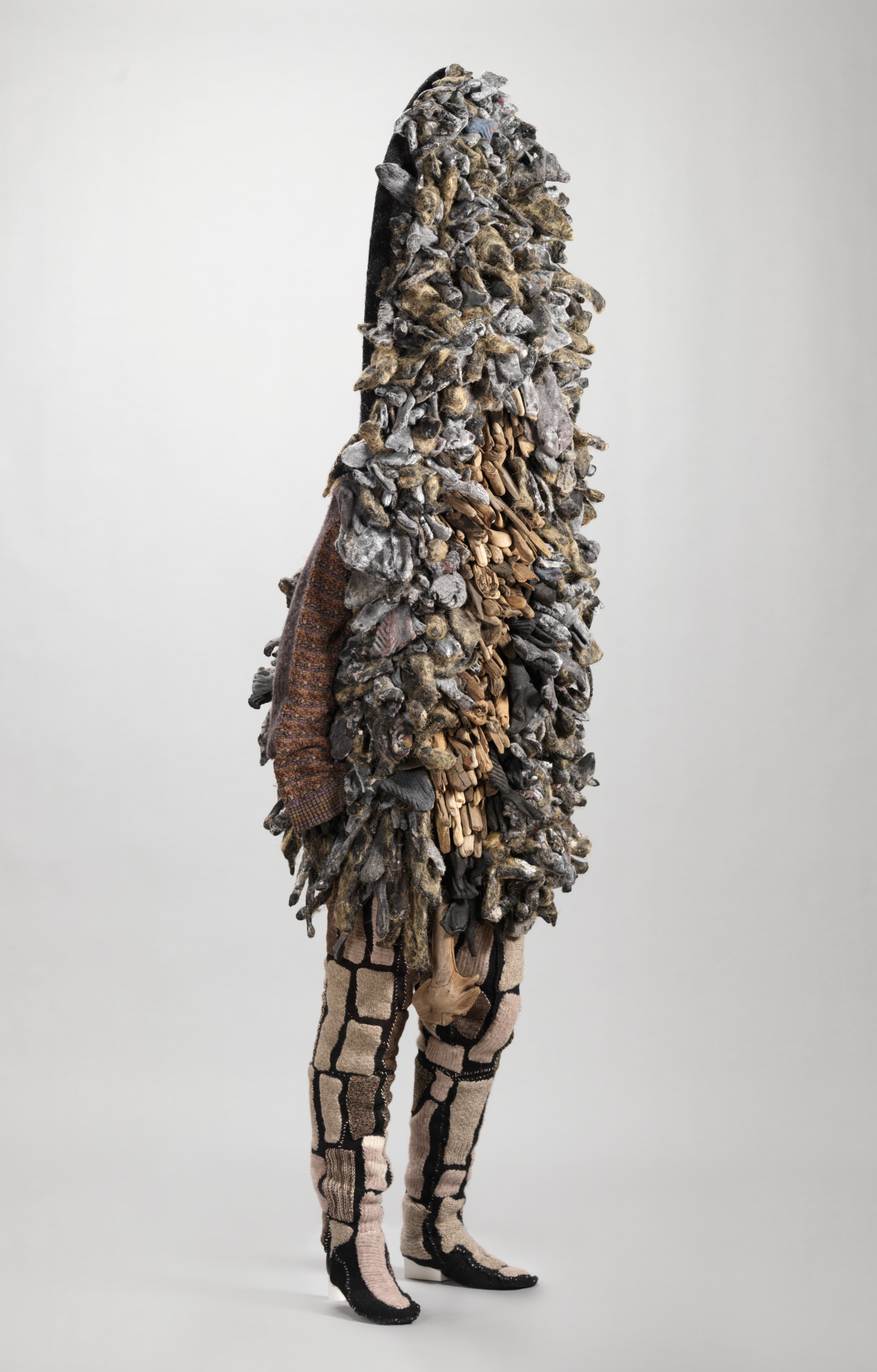 Side view of full-body sculptural garment in earthy tones engulfing the upper body in dense glove or finger-like forms with a sweater sleeve hanging out and legs clad in patchwork.