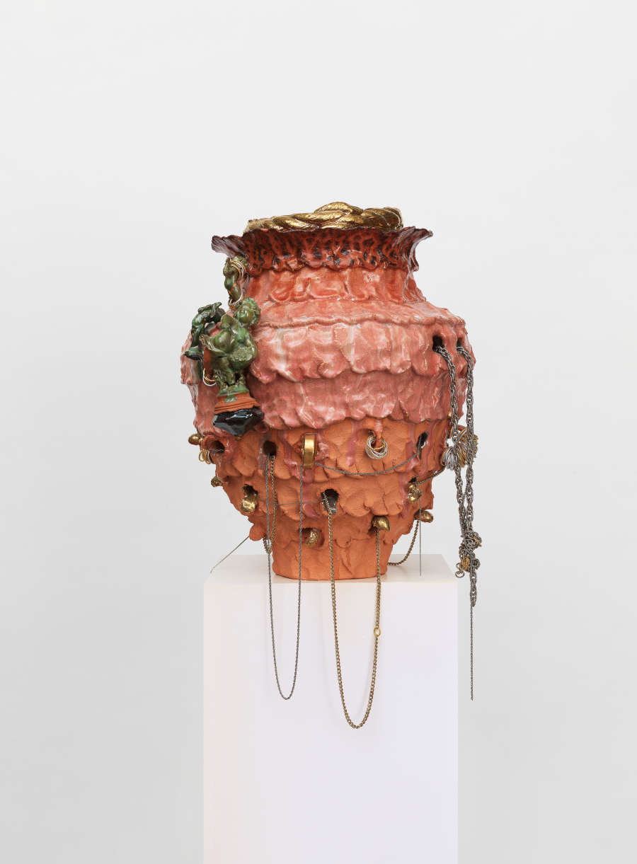 Coral-colored ceramic urn with a rough, textured surface and hints of gold and foliage, the lower half punctuated by small holes from which delicate metallic chains drape and dangle loosely.