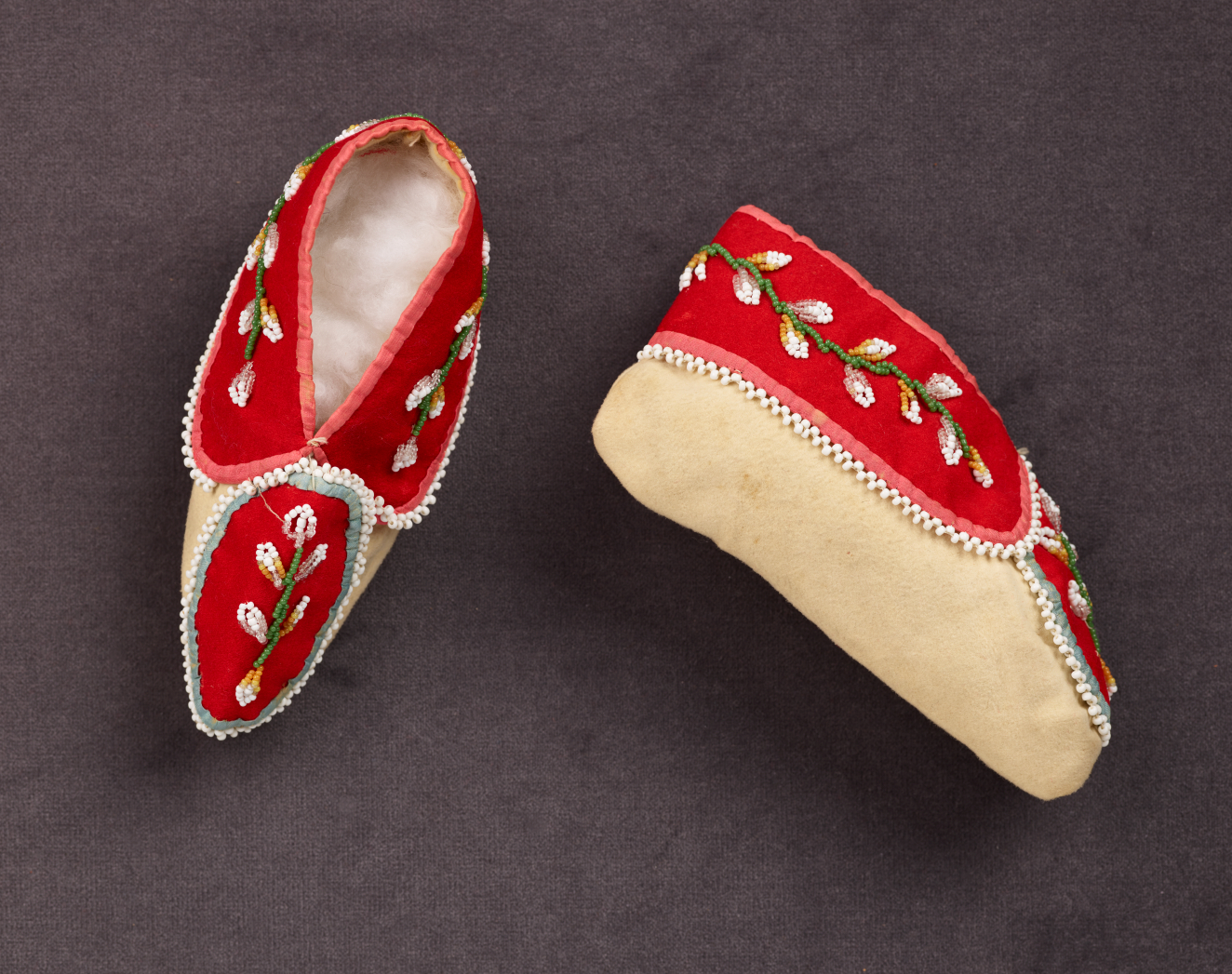 Pair of red moccasins featuring white and green floral embroidery as well as tan soles. One moccasin sits right-side-up on the gray surface while the other is on its side.