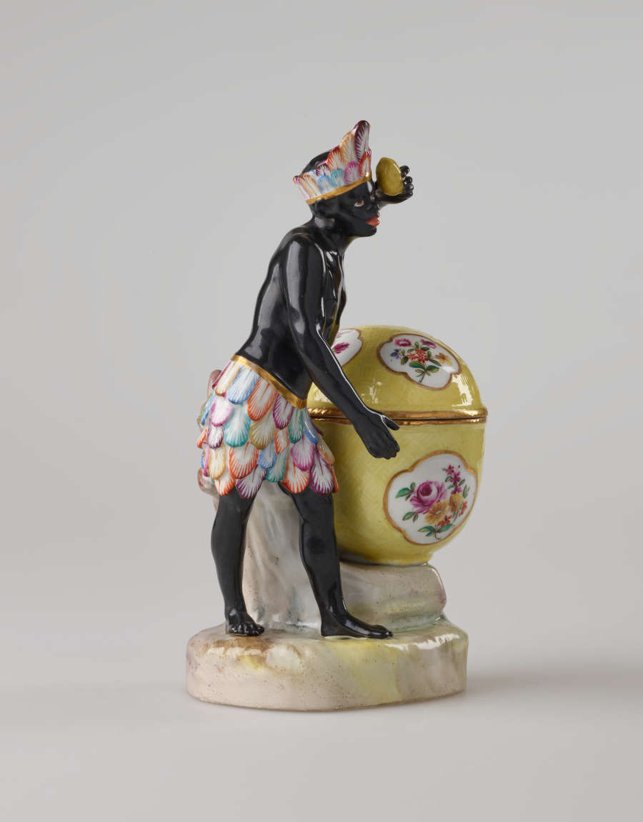 A sculptural figure with black skin, a colorful headdress and skirt. This figure is standing next to a bright yellow covered sugar basket with floral decorations.