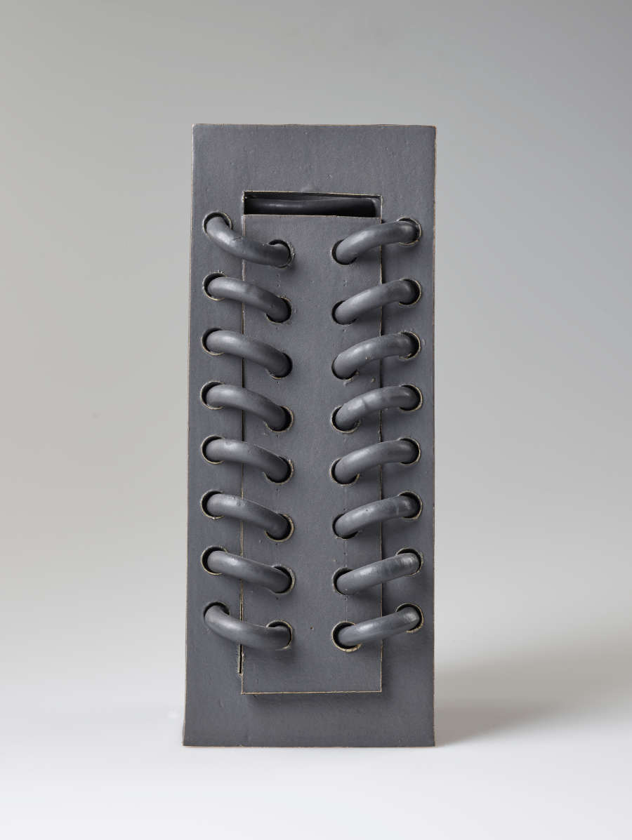 Straight-on front view of a rectangular gray ceramic sculpture featuring a row of horizontal loops threaded and protruding from the surface. The is a smaller rectangle carved into the center.