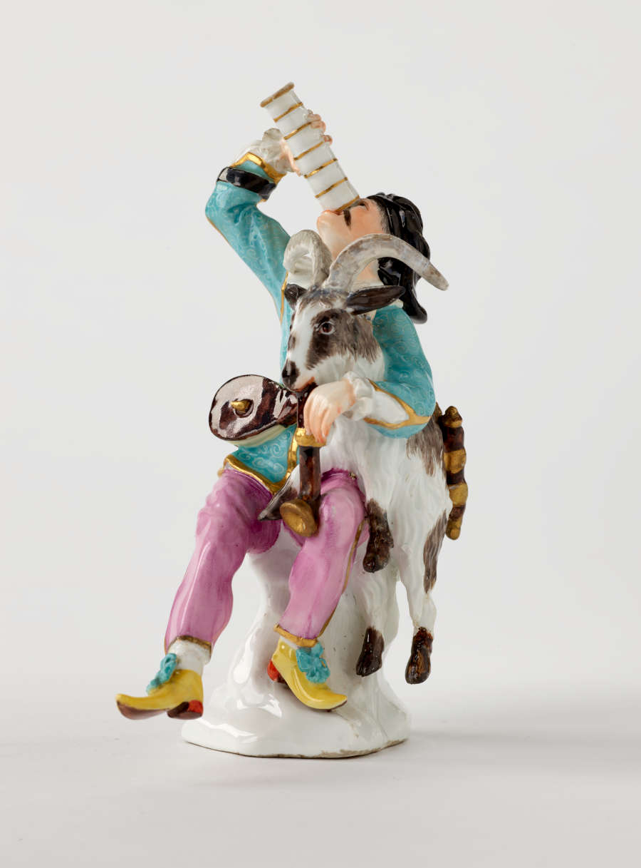 Sculpture of a light skinned man with a goat on his lap. He holds a dark brown instrument in one hand and drinks from a vessel in the other.