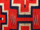 Closeup of the center of a red woven cloth, showing a navy blue and cream bordered cross design in between two navy blue and cream zig zag patterns. 