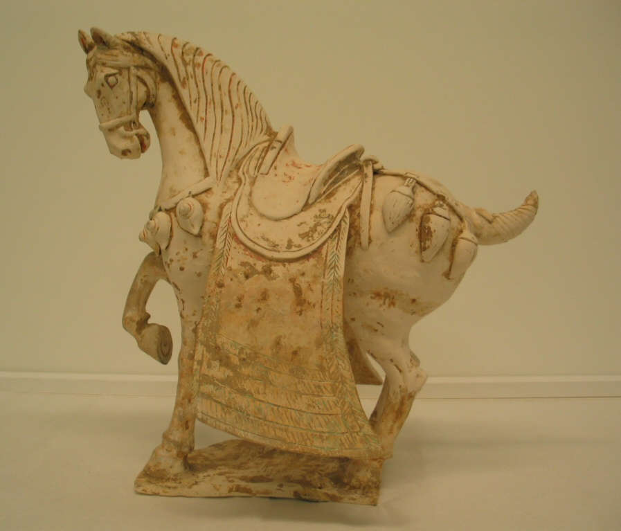 Another side-view of a white ceramic sculpture of a saddled, ornamented horse with one foot raised, a short wound tail and heavy mane. It stands on a thin connected base.