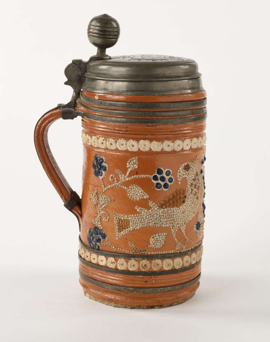 An orange stein with a metal lid that is hinged to the handle. The white and blue decorations consist of dots and are primarily depicting floral imagery.
