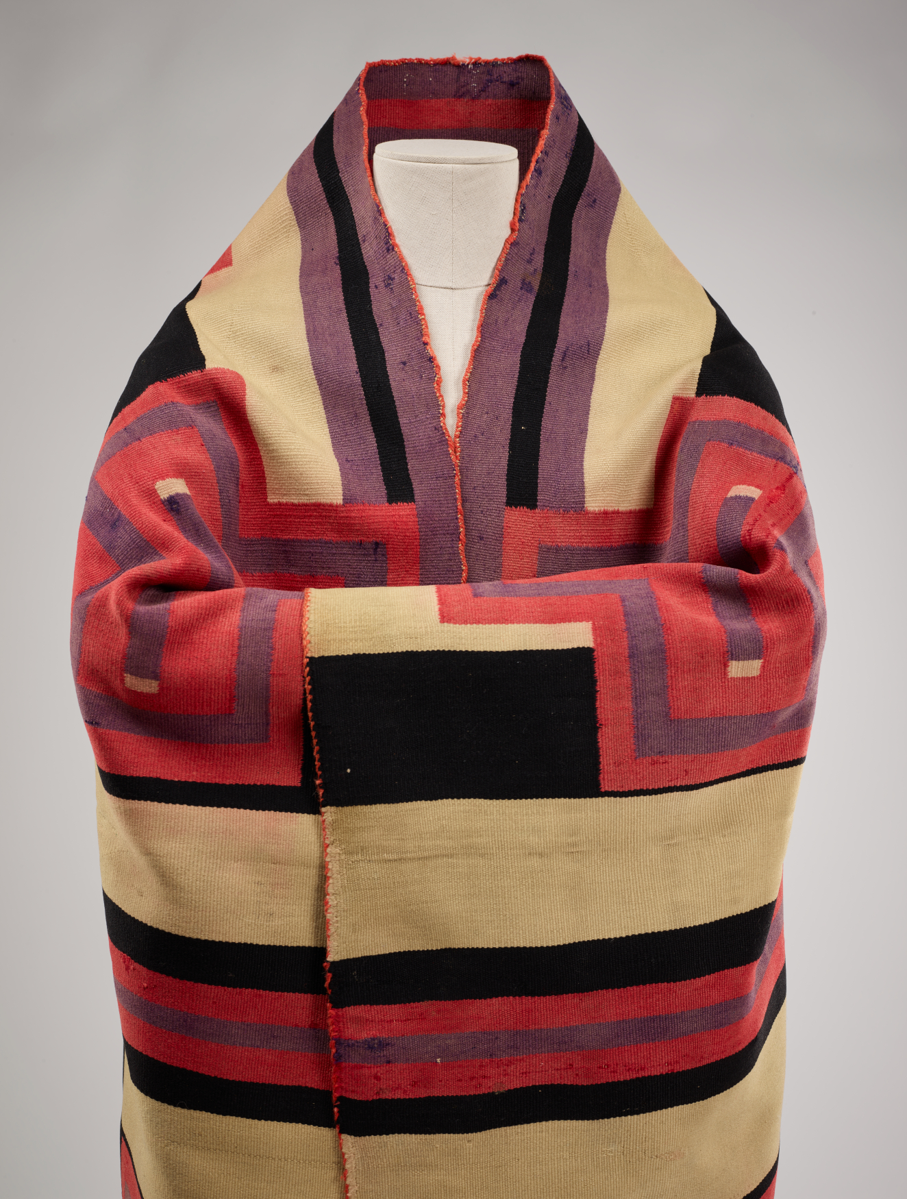 Closeup of a black and cream striped woven robe wrapped around a mannequin with folded arms. Three thin purple and black stripes line the hem of the robe's deep collar. 