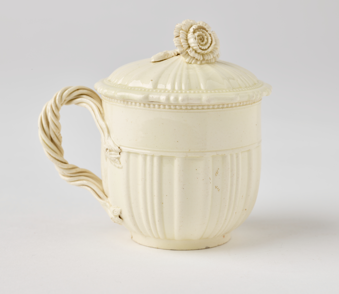  A cream-colored cup with a sculptural swirled handle, and lid with sculpted finial.