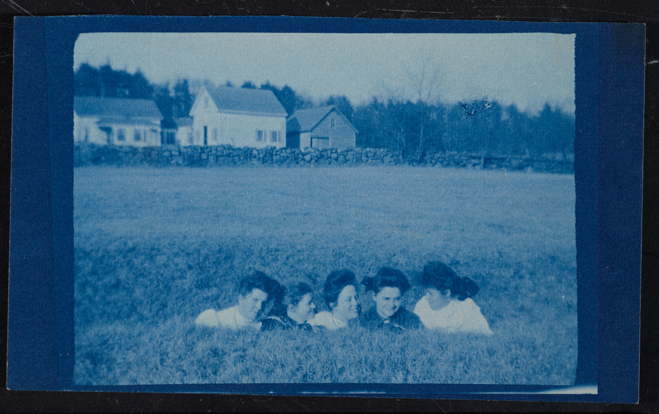 Blue-toned vintage snapshot of five smiling young light-skinned women. They are in the middle of a field, and only their heads and shoulders are visible. White houses rise behind them. 