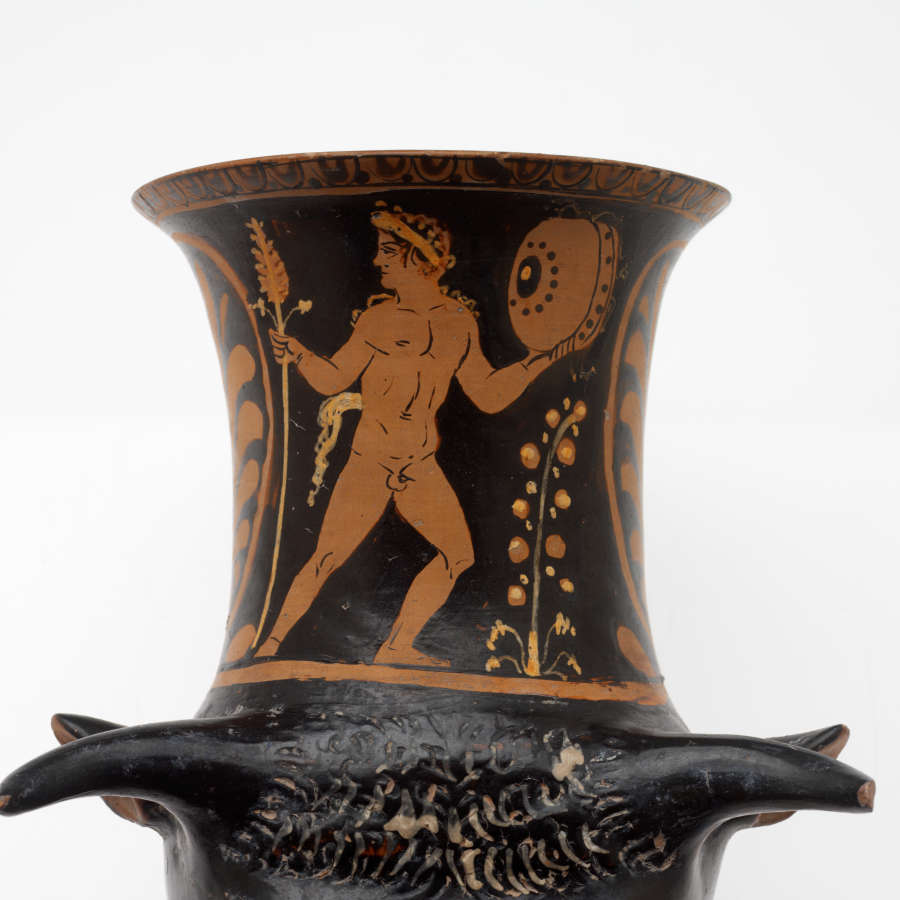 Detail view of the black and terracotta cup’s neck, showing that it is fluted with illustrations of nude men holding grains and a shield next to floral motifs. 