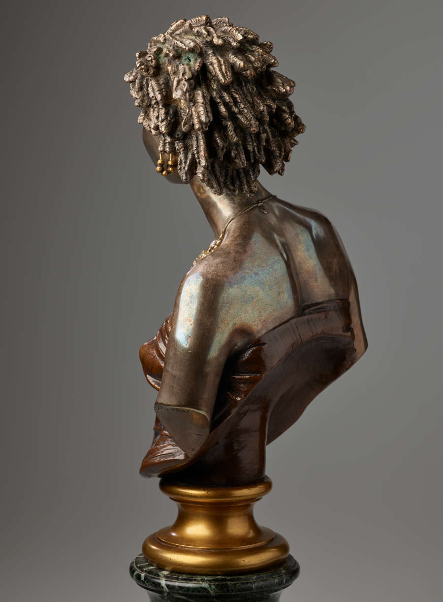 Rear-left angle view of a bronze bust highlighting hair in twists and sculpted, curved shoulder blades exposed above a dark strapless top cut off diagonally and mounted on a gold base.