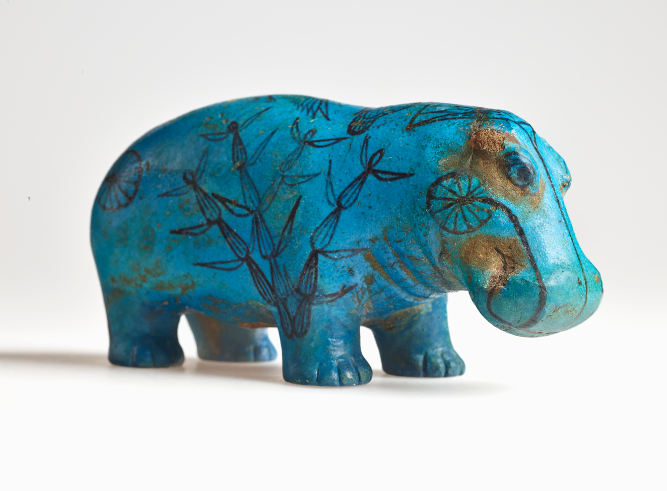 Side view of a bright turquoise hippopotamus decorated with black linework depicting reeds and papyrus fronds, its surface marked by splotches and lines showing the clay underneath the turquoise glaze.