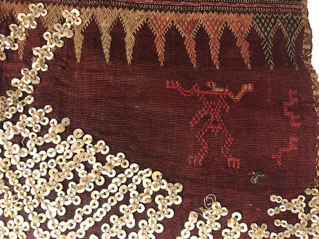 Closeup of a deep red woven fabric with geometric embroidery depicting a triangle border and a humanoid figure in red. Along the bottom are cascading reflective sequins.