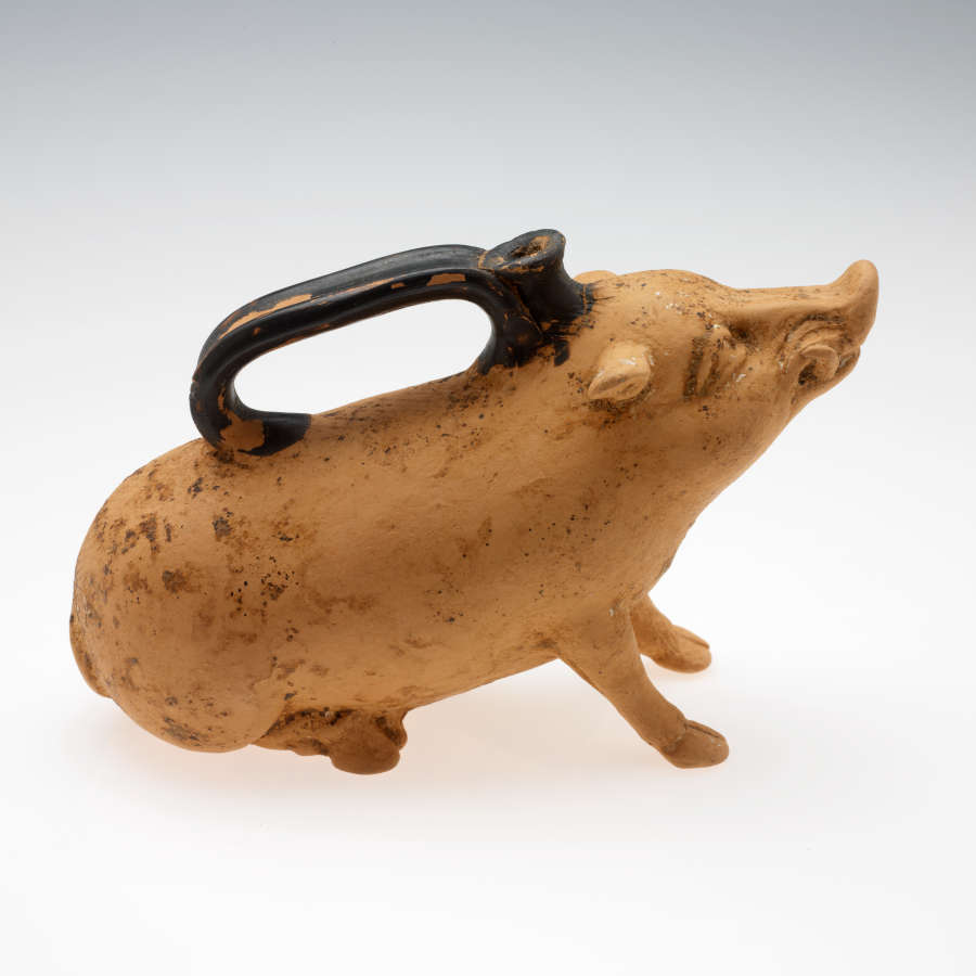 Side-view of a terracotta container shaped as a seated boar with an upturned snout looking upwards. Along its back is a chipped long black handle with a narrow connected mouth.