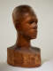 Three quarter view of a textured, but glossy, wooden-carved bust depicting a male figure with afrocentric facial features. The expression is cast in shadow, highlighting the side of the head.