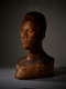 Three quarter view of a wooden bust depicting the head of a male figure with afrocentric facial features. He has a somber expression showcased half in light, half in shadow.