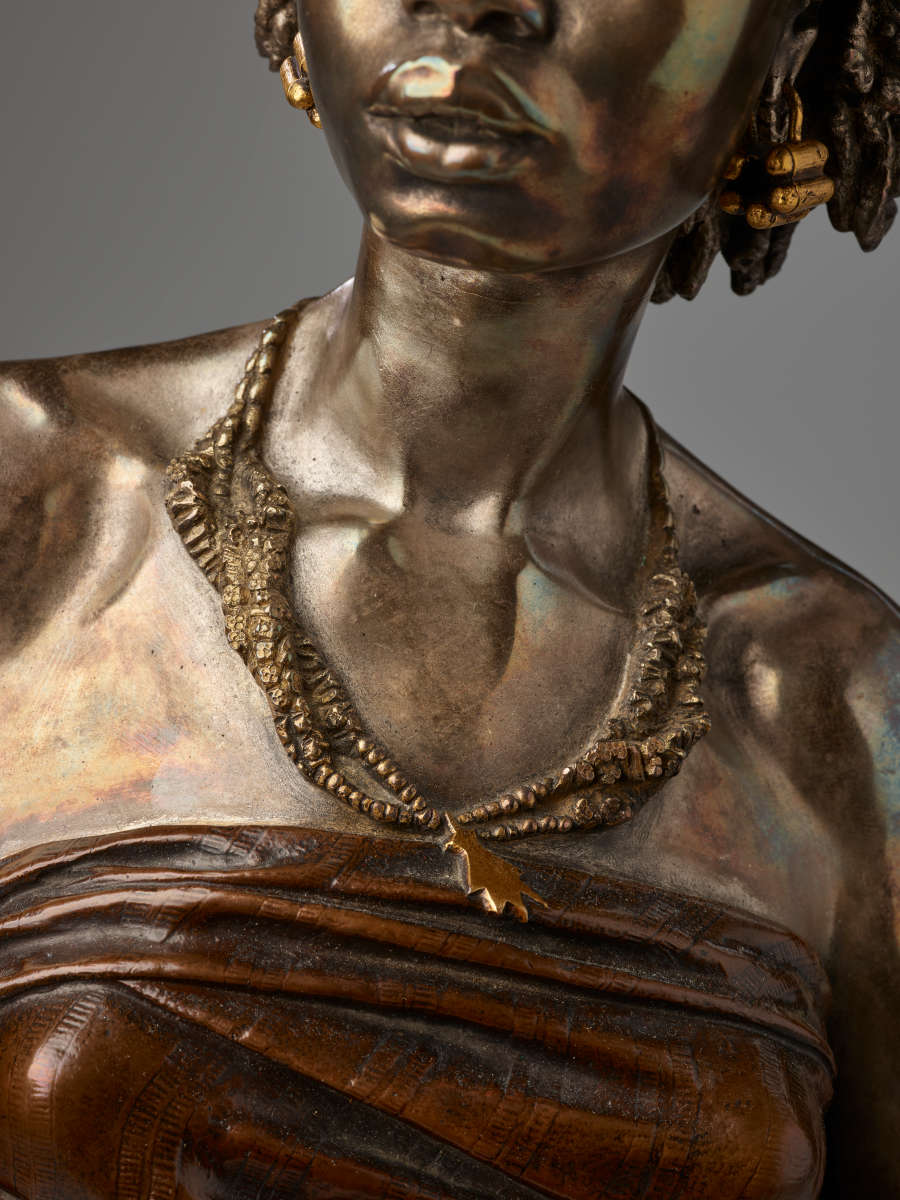 Close-up of bronze chest with twisted beaded necklaces with leaf charm resting on exposed collarbones above a dark strapless tightly-wrapped cloth top.