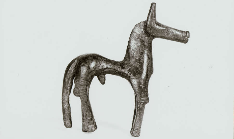 A monochrome side-view of a tarnished bronze horse figurine with an exaggeratedly thin torso, thick neck, long legs and tail and long narrow snout.