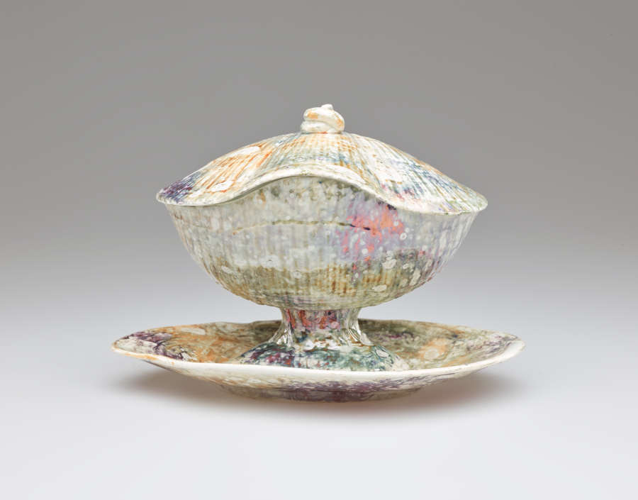 A lidded vessel on an organically shaped tray. A small, swirled finial is on the lid. The body of the vessel has ridges covered with a mottled, multi-colored glaze.