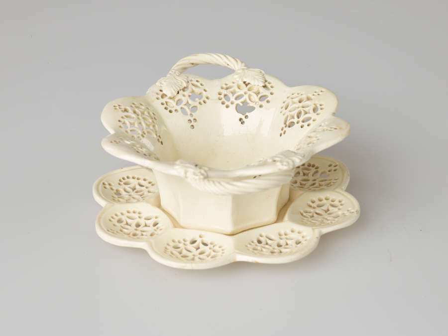A cream-colored dish with two swirled handles and pierced decorations. Dish sits on a matching tray.