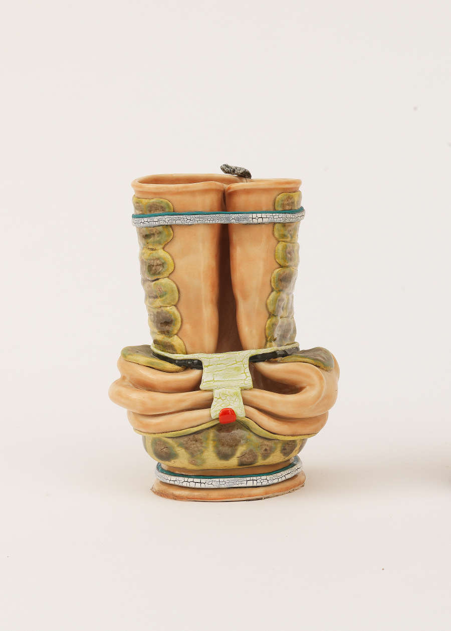A small ceramic vessel of intertwined hands gripping a scroll decorated with green patterning resembling snake skin and a tiny snake resting on the top rim. 