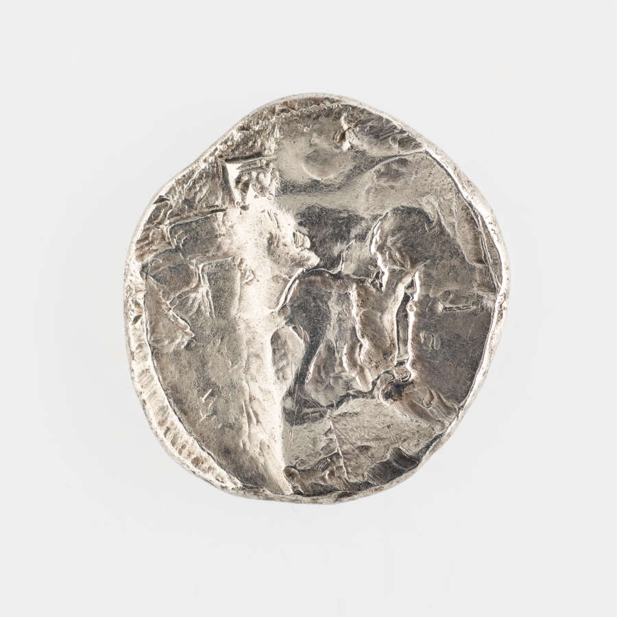 Flip-side of the silver coin, showing that its surface is uneven in texture and color. The coin’s round edge is uneven.