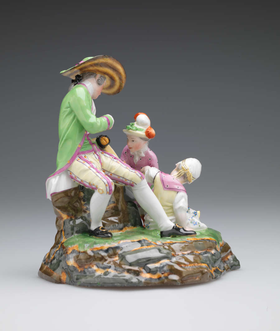 A sculpture with three figures in elaborate historical clothing. A seated light-skinned figure holds a roll of maps while one child examines. A second child sits near them.