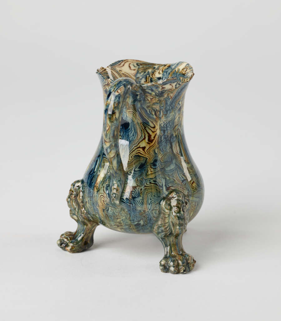 A small vessel with a cream, blue, and green mottled design. Three sculptural feet are shaped like lion’s paws with lion heads attached to body of vessel.