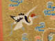Embroidered textile featuring a red-capped crane with outstretched black and white wings, set against a gold background with swirling blue and pink clouds. 